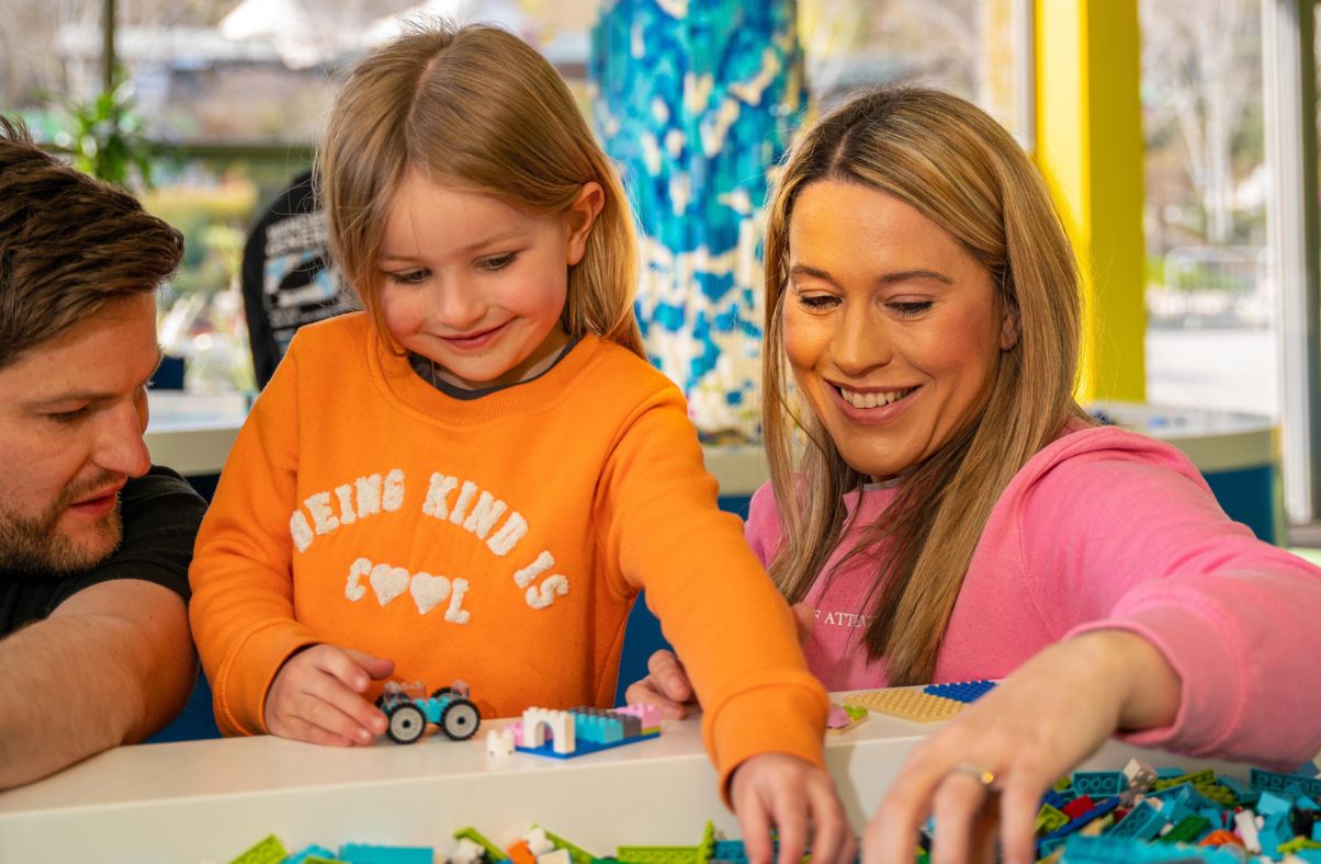 A young girl and two adults playing together with colourful LEGO bricks at a building table.