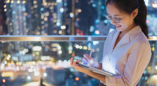 Smiling businesswoman using a digital tablet at night with a city skyline in the background, symbolizing engaged learning and AI readiness in the workplace.