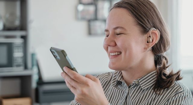 Person engaging with content on a smartphone, smiling, wearing a hearing aid in a comfortable work setting.